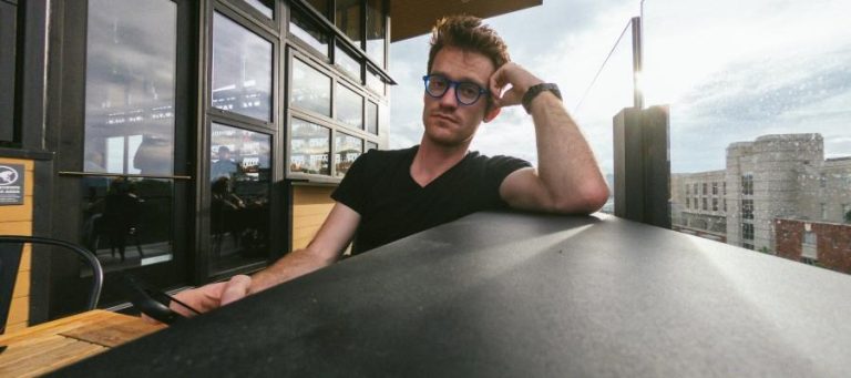 Man wearing glasses sits at outdoor table, contemplating.
