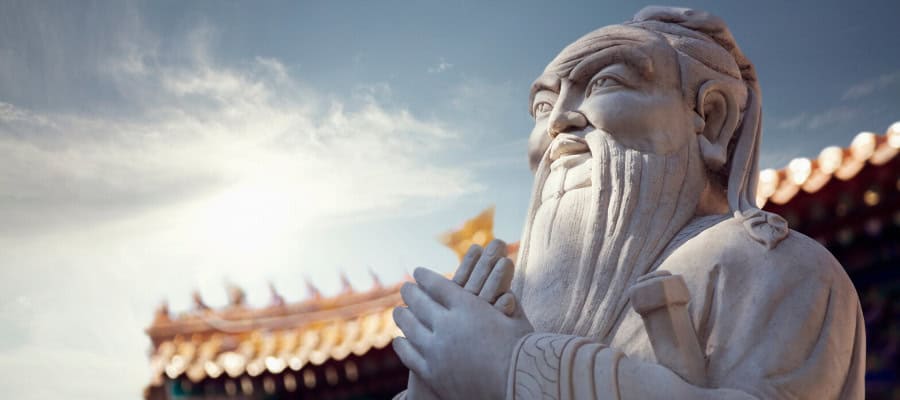 Stone statue of Confucius, hands clasped, against a temple backdrop.