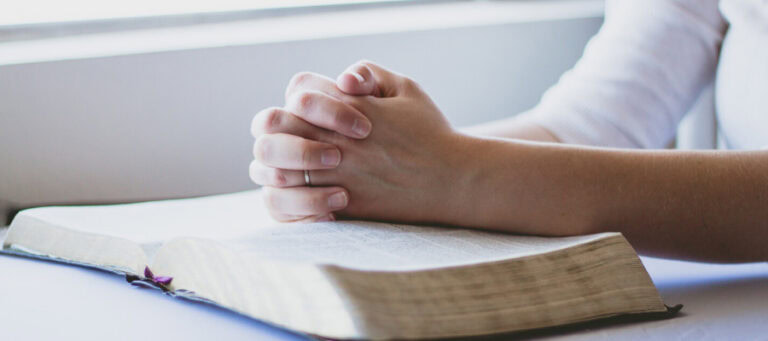 Woman's hands clasped in prayer atop an open Bible.