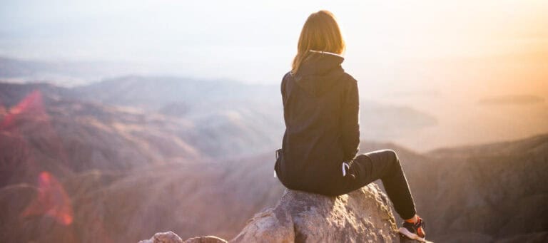 Woman sitting on a rock overlooking a vast mountain range at sunset, contemplating God, design, and science.