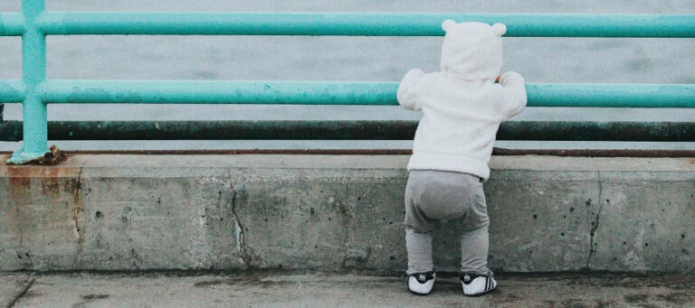 Toddler in hooded jacket looks over a railing.