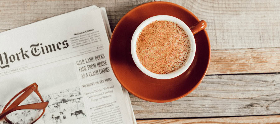 A cup of coffee sits on a saucer next to a newspaper with the headline "New York Times".