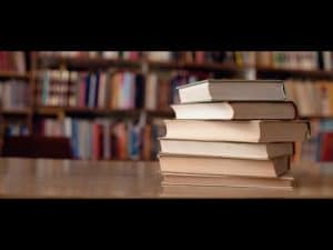 Stack of books on a table in front of bookshelves. Language of fiction.