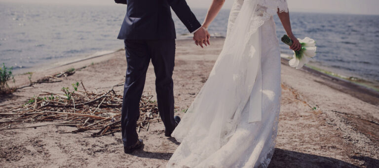 A bride and groom hold hands, walking away from the camera on a beach.