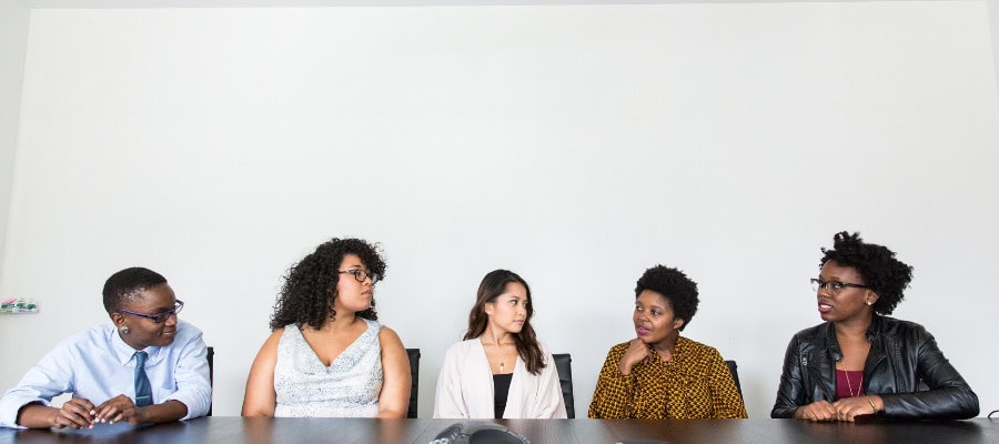 Five diverse professionals sit at a conference table, engaged in discussion.