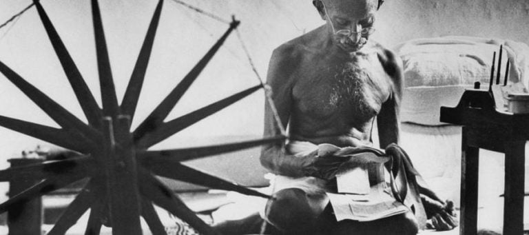 Mahatma Gandhi, seated, spinning yarn on a charkha, appears contemplative.