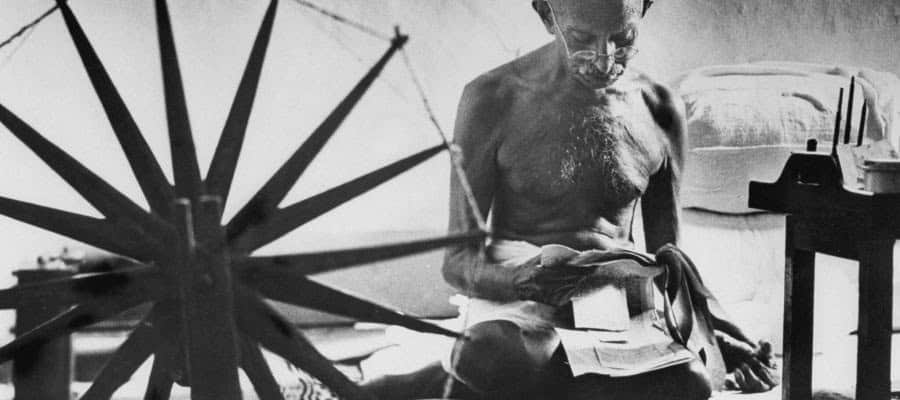 Mahatma Gandhi, seated, spinning yarn on a charkha, appears contemplative.