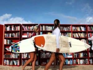 Two women carrying surfboards near a red bookcase filled with summer reading books.
