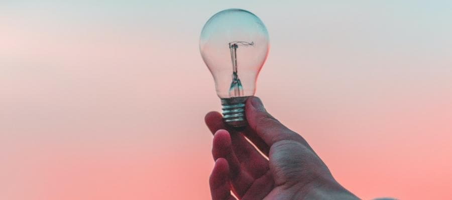 Hand holding a lightbulb against a pink and blue sky.