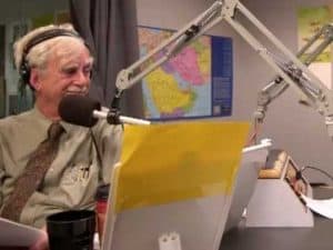 Older man in a shirt and tie speaks into a microphone during a radio broadcast.