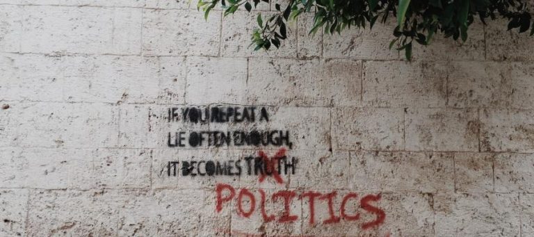 Graffiti on a stone wall reads: "If you repeat a lie often enough, it becomes truth" crossed out, with "POLITICS" below.