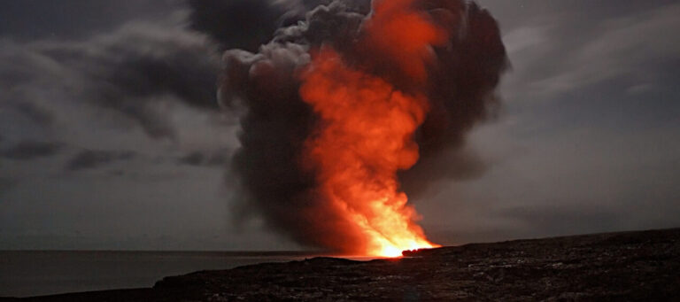 Fiery volcanic eruption at night, billowing dark smoke and bright orange lava.