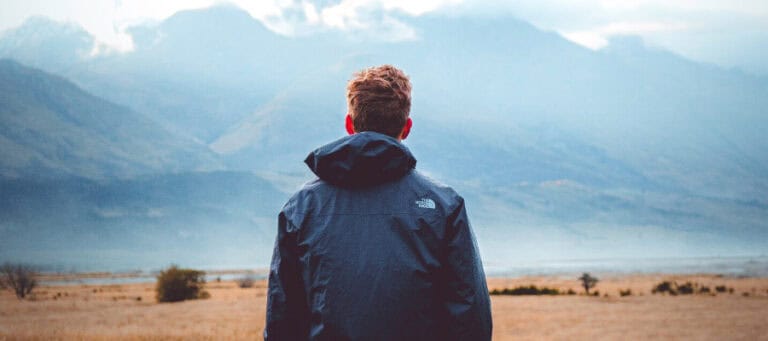 Man in hooded jacket contemplates vast mountain landscape.