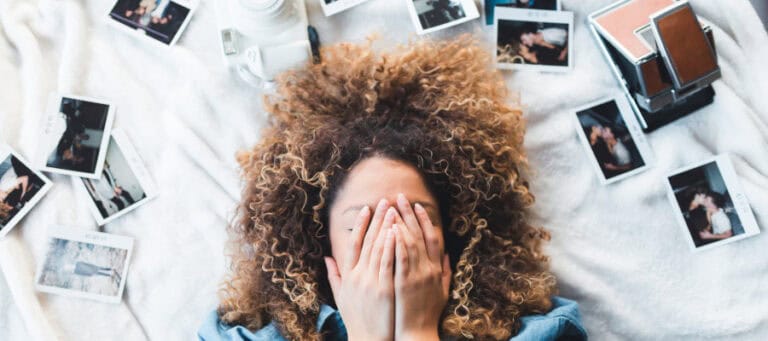 Woman with curly hair covering her face with hands, surrounded by photos.
