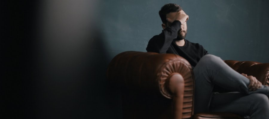 Man sits on a leather couch, hand on forehead, appearing stressed.
