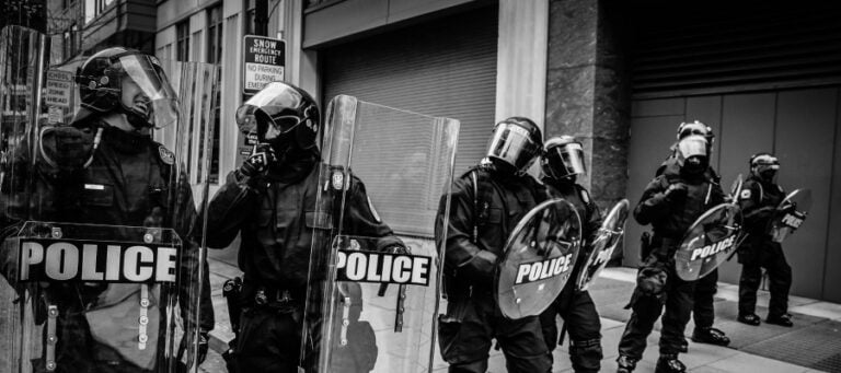 Police officers in riot gear stand in formation, holding shields marked "POLICE.
