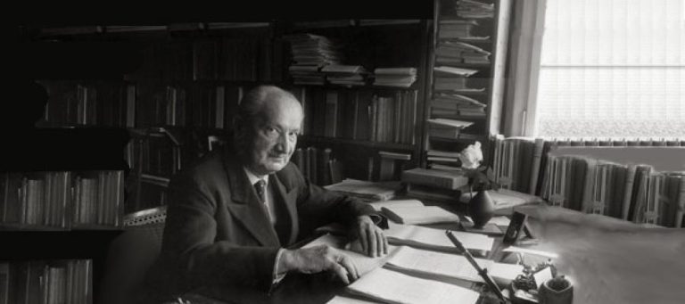 Black and white photo of elderly man seated at desk surrounded by books, contemplating.