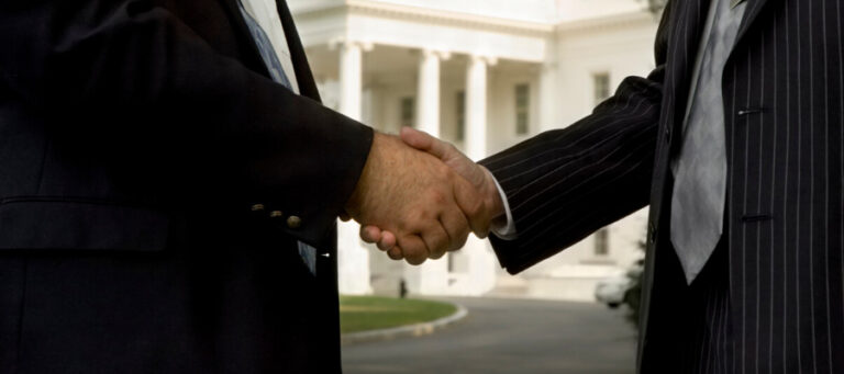 Two men in suits shaking hands in front of the White House.