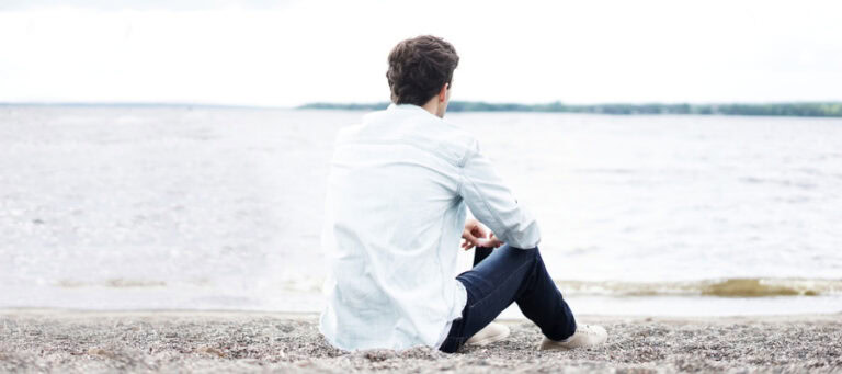 Man sitting on beach, looking out at water. Contemplative pose suggests a safe space.