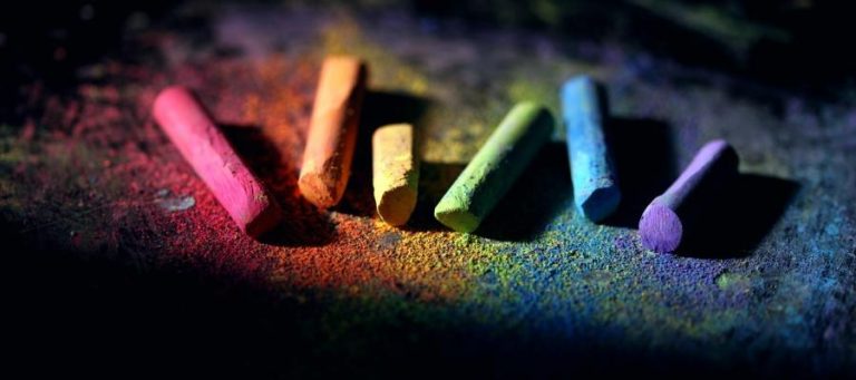 Rainbow-colored chalk pieces on a dark surface, surrounded by colorful chalk dust.