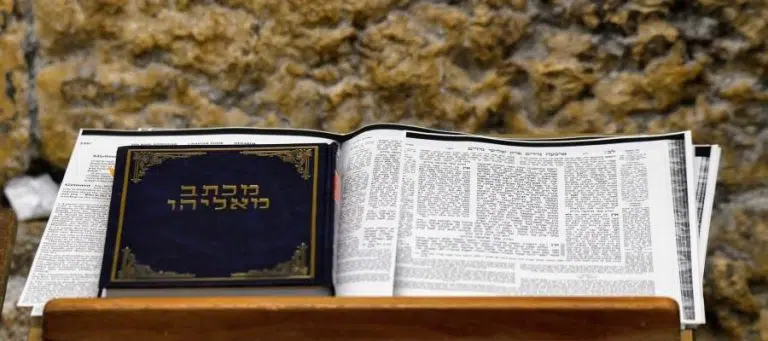 Open Hebrew prayer book on wooden stand against ancient stone wall. The book cover displays Hebrew text.