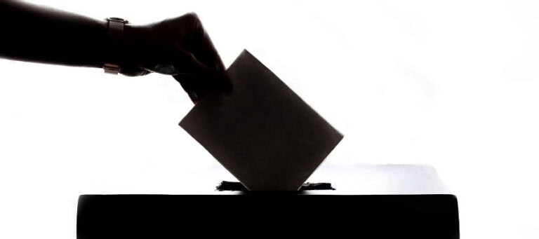 Hand dropping ballot into a ballot box, exercising the right to vote. Dewey's Democracy.