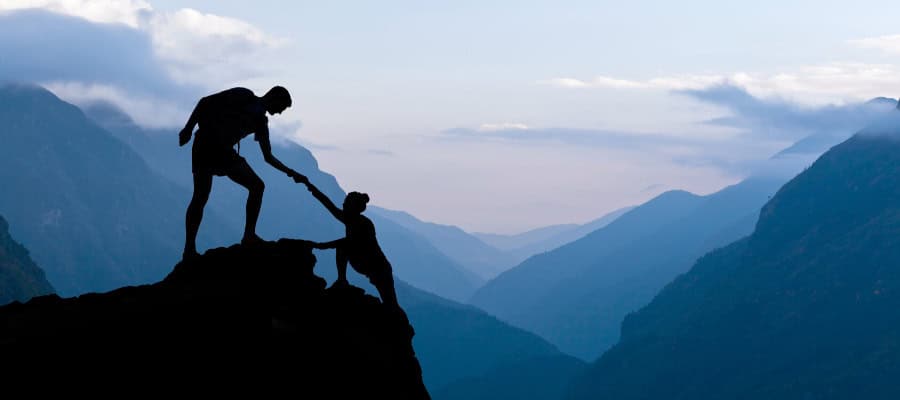 Silhouette of two hikers helping each other climb a mountain.