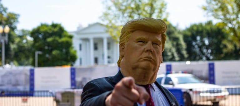A person wearing a Donald Trump mask points at the viewer near the White House.