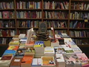 Stack of books on a table, surrounded by bookshelves filled with various titles. Summer reading list.
