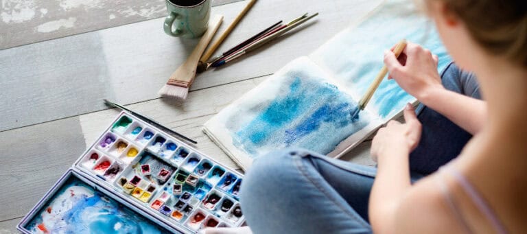 Woman painting a blue watercolor landscape. Art supplies including brushes and paints are visible.