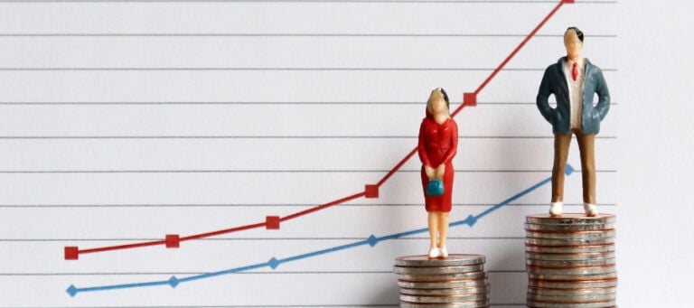 Miniature figures stand atop stacks of coins illustrating a pay gap.