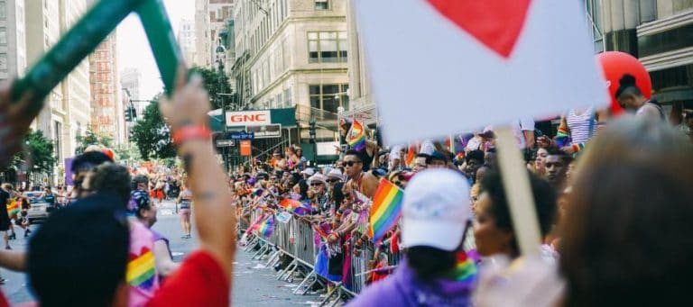Crowd at a Pride parade, holding rainbow flags and signs.