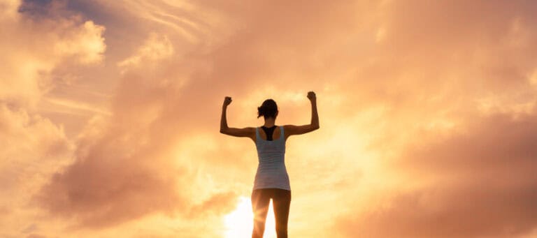 Woman silhouetted against sunset, arms raised in victory pose.