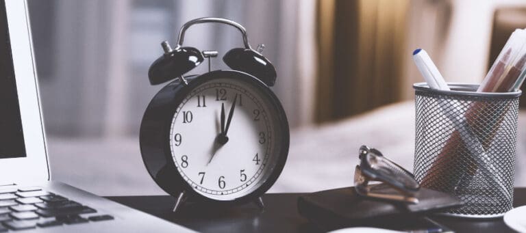 Black alarm clock showing almost noon, next to a laptop and glasses.