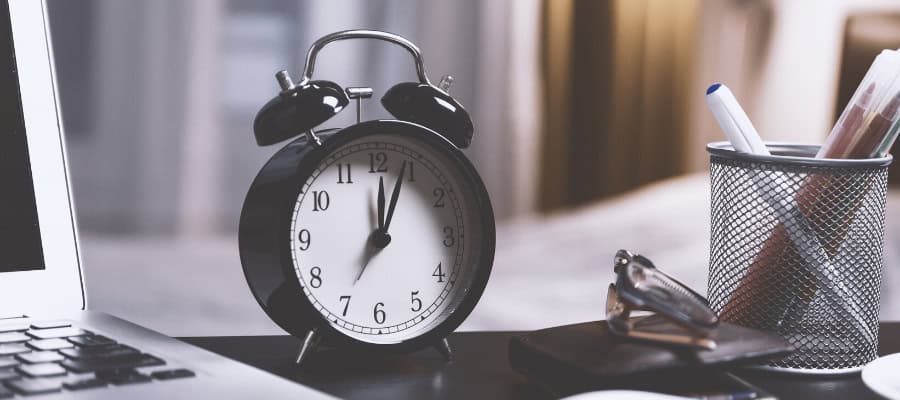 Black alarm clock showing almost noon, next to a laptop and glasses.