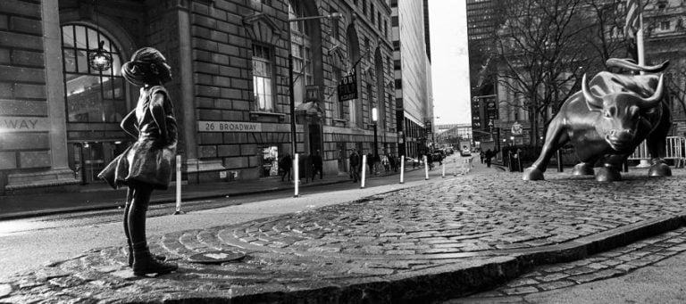 Black and white photo of the Fearless Girl statue facing the Charging Bull statue in New York City's financial district.