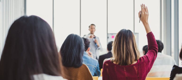 Attentive audience listening to a speaker at a seminar; one person raises their hand to ask a question.