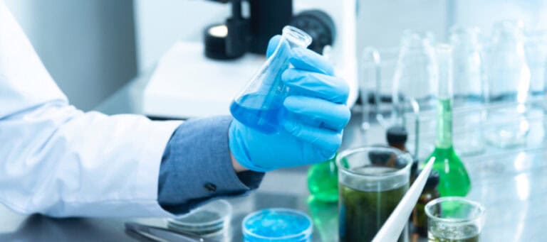 Scientist's gloved hand holding a flask of blue liquid in a lab.