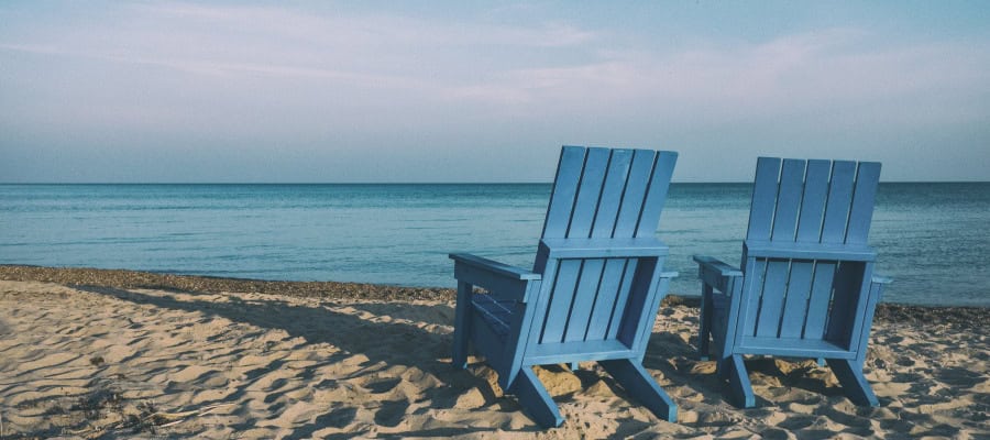 Two blue Adirondack chairs on a sandy beach facing the ocean.
