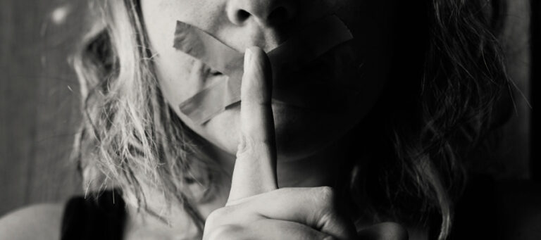 Close-up of a woman's mouth covered with tape, finger to lips.