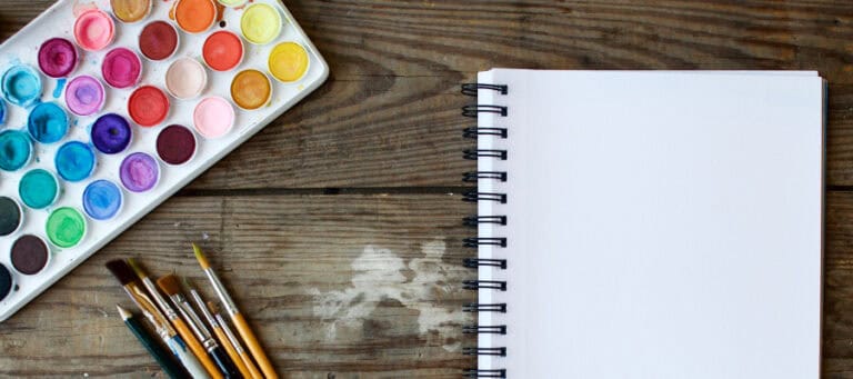 Watercolor paints, paintbrushes, and a blank spiral notebook sit on a wooden surface.