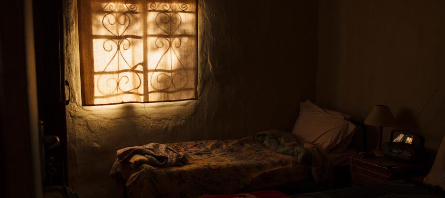 Dark bedroom with a single bed, a small table, and a vintage radio. Sunlight streams through a window with an ornate metal grille.