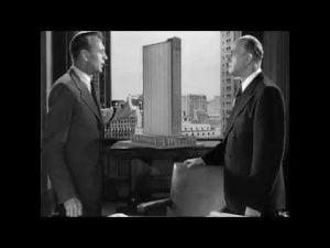 Two men discuss a building model, debating the political implications of architecture.