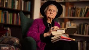Mary Midgley, author, sits in a chair surrounded by books, holding a stack of books.
