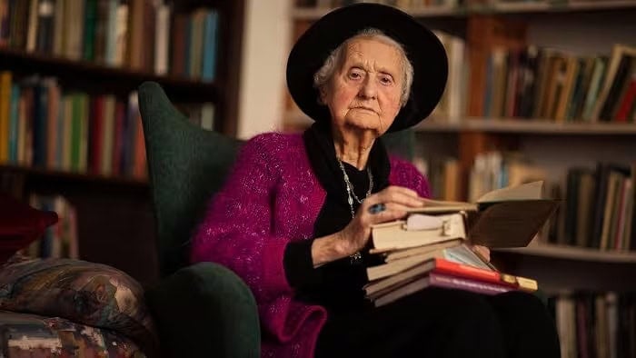 Mary Midgley, author, sits in a chair surrounded by books, holding a stack of books.