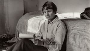 Black and white photo of Iris Murdoch, author, sitting on a bed, holding papers.