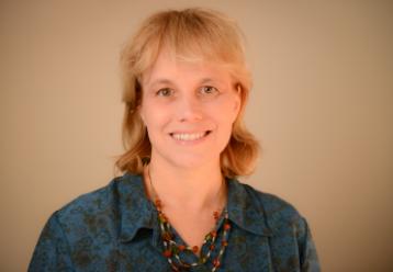 Headshot of a smiling woman with blonde hair, wearing a teal patterned shirt and necklace.