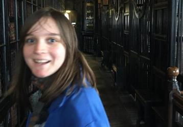 Happy young woman in a blue jacket, exploring a dark, book-lined corridor.
