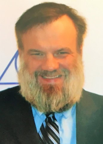 A man with a beard, wearing a suit and tie, smiling warmly.