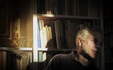 Man sits in a dimly lit room, surrounded by bookshelves.